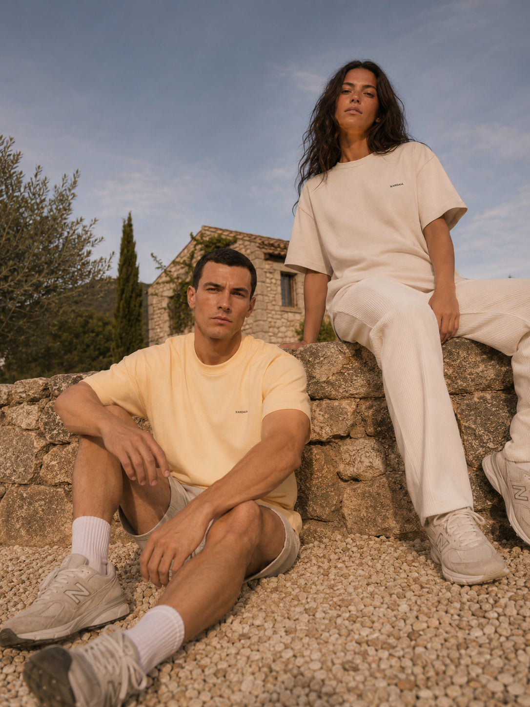 Homme et femme portant des T-shirts oversize Kardaia en coton biologique, coloris Lin clair et Sable doux, dans un décor méditerranéen.