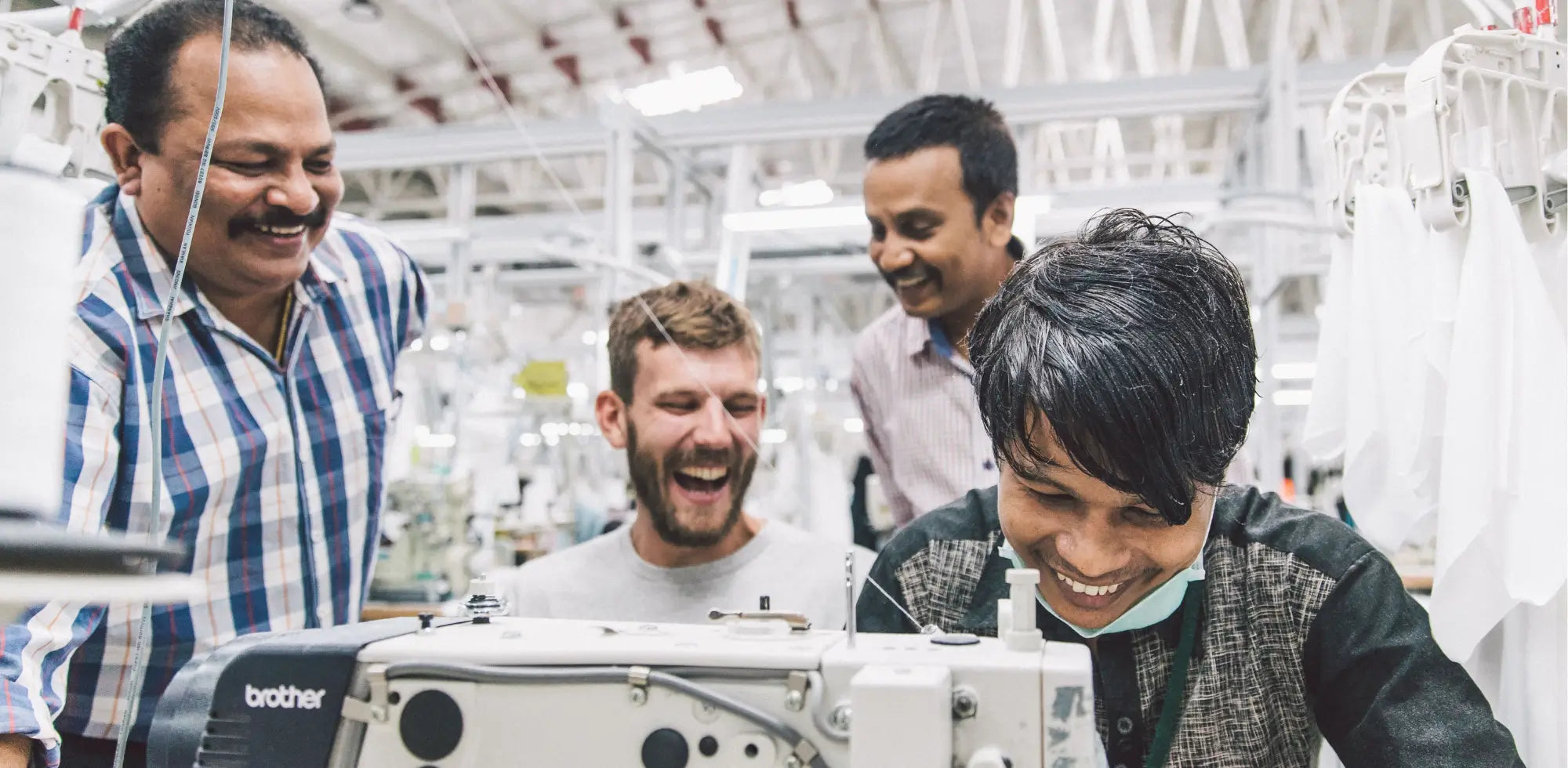 Couturier souriant à sa machine à coudre, entouré de son équipe dans un atelier partenaire en Inde