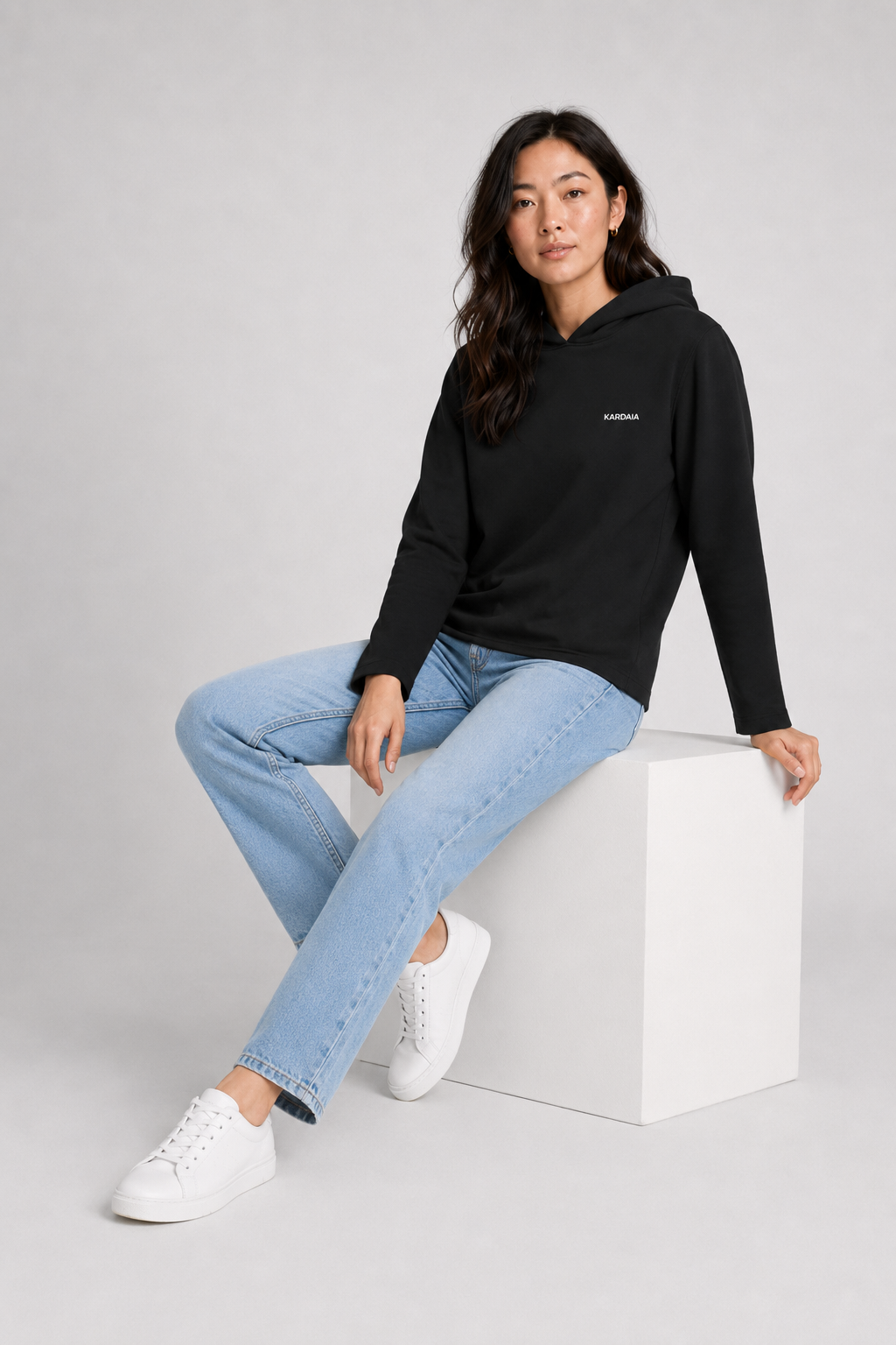 Femme portant un sweat à capuche noir en coton biologique avec un jean bleu assise sur un cube