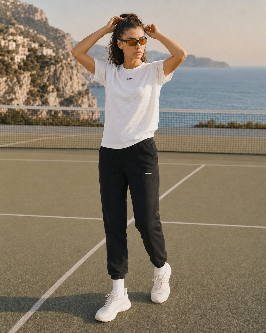 Tenue femme Kardaia composée d’un t-shirt blanc en coton biologique et d’un pantalon de survêtement noir assorti. Photographie en pied sur un court de tennis avec les falaises d’Èze et le littoral méditerranéen en arrière-plan.