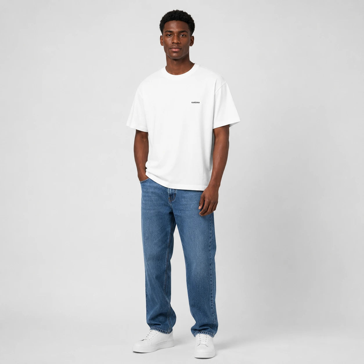 T-shirt homme Kardaia blanc en coton biologique, présenté dans une coupe classique droite et confortable. Porté avec un jean bleu et des baskets blanches sur un fond de studio gris clair.