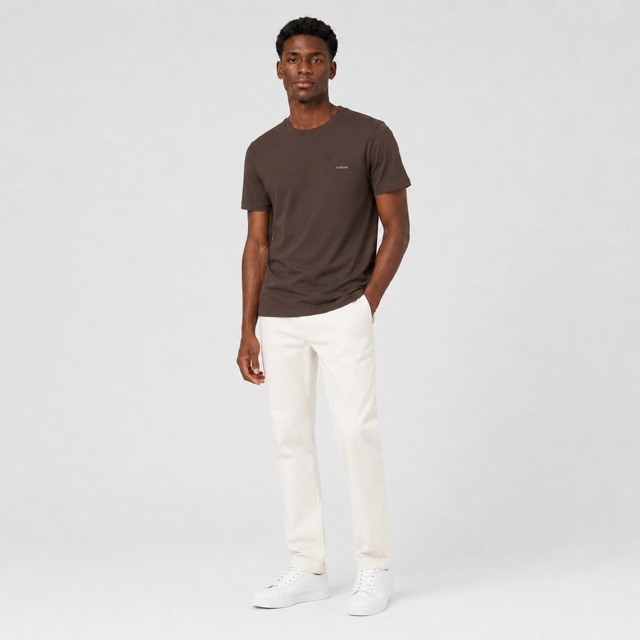 T-shirt homme Kardaia marron en coton biologique, conçu dans une coupe classique légèrement ajustée. La photographie en pied met en valeur son tombé droit, ses manches courtes et sa teinte brune avec un pantalon blanc cassé.