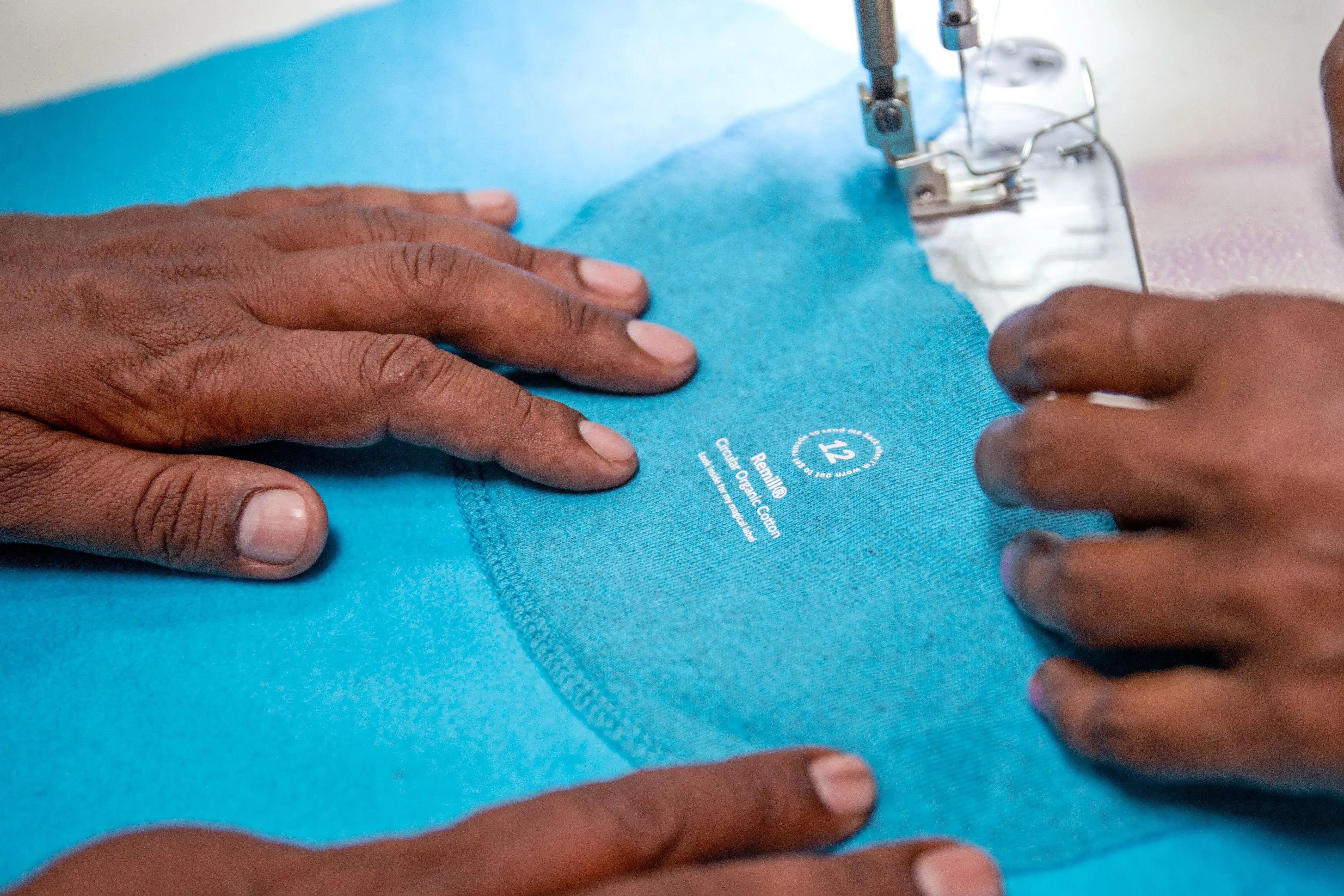 Mains d’artisan cousant une étiquette en coton bio Remill sur un vêtement bleu, démarche textile circulaire