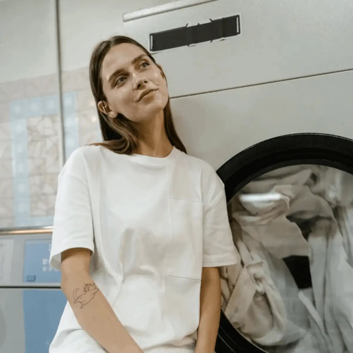 Femme en t-shirt blanc assise devant une machine à laver, ambiance douce et posée