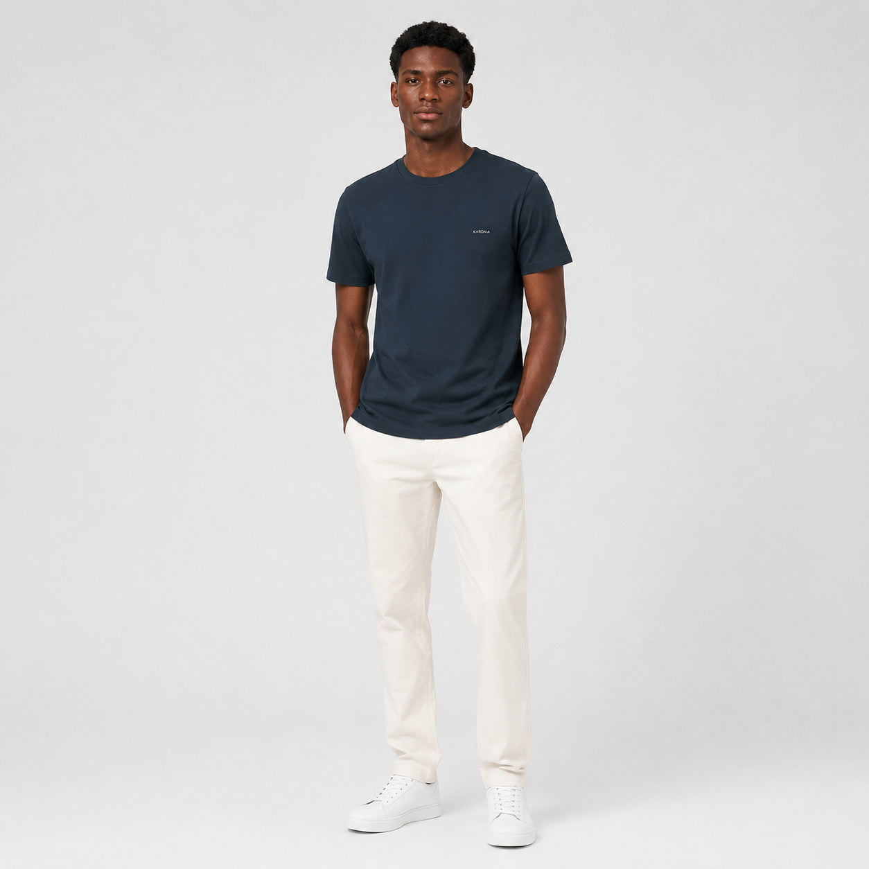 T-shirt homme Kardaia bleu marine en coton biologique, doté d’une coupe classique droite et d’un col rond. Présenté avec un pantalon écru et des baskets blanches dans un décor de studio minimaliste.