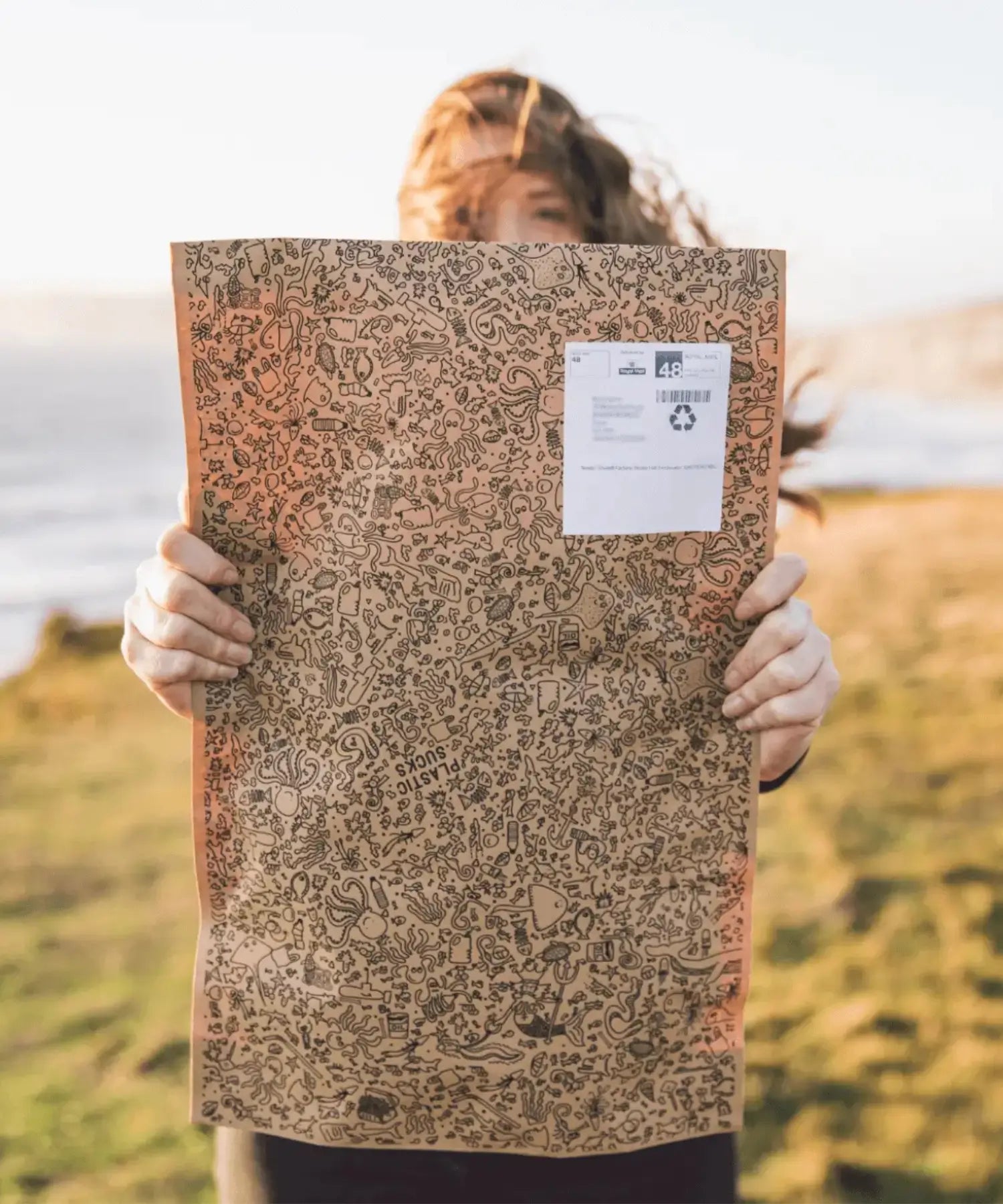 Femme tenant un emballage recyclable illustré, en bord de mer, solution d’envoi sans plastique